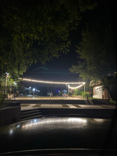 This is a nighttime photograph taken outdoors in Gorna Malina, Bulgaria. The image captures a pathway leading towards a stage or performance area, illuminated by strings of lights and various lampposts. The foreground is dominated by the glossy, dark surface of what appears to be a car's hood, reflecting the overhead lights. Beyond this, a series of wide, shallow steps lead up to a paved plaza. A low, stone-faced wall borders the left side of the plaza, with a metal enclosure containing some decorative elements behind it.
In the middle ground, a long string of warm, yellow lights stretches across the scene, suspended between poles. Several lampposts with white lights are also visible, casting a glow on the ground. Further back, a small carousel with several figures, likely children's rides, is lit up. Trees with dense foliage frame the upper edges of the photograph, their leaves illuminated by the artificial light. A building with a prominent window is visible on the right side of the image, constructed with brick. In the distant background, a faint, arched structure with more lights is visible against the dark sky. No people are clearly discernible in the image, though the presence of the carousel suggests a public space that may be frequented by families. The overall impression is one of a quiet, public park or gathering space at night.