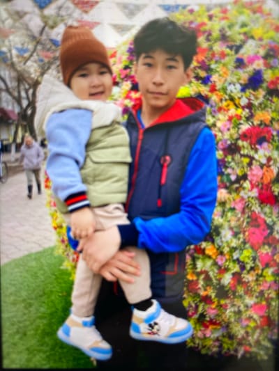 The image shows two young individuals, likely brothers, with the elder holding the younger in his arms. The elder boy, with dark hair, is wearing a blue and dark gray vest over a blue long-sleeved shirt with a red collar. The younger child, a toddler, is wearing a brown knit hat, a light-colored vest over a light blue garment, and beige pants. The toddler's shoes are white and blue with a Mickey Mouse design. They are standing in front of a vibrant, colorful floral arrangement. In the background, on the left side of the image, a person in a light-colored hoodie and pants is walking away from the camera. The ground in the foreground appears to be a combination of green artificial turf and patterned paving stones. The overall setting suggests an outdoor public space, possibly a park or a festival, within the Бескөл ауылдық округі, Kazakhstan.