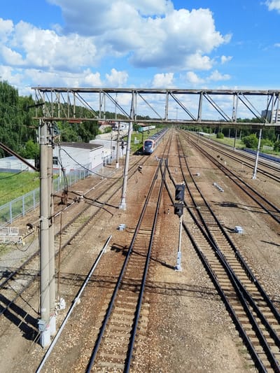 The media file shows a wide view of a train yard with multiple tracks under a bright blue sky with fluffy white clouds. The scene is set in Monino, Russia, as indicated by the location context. The time of day appears to be midday, given the strong sunlight casting shadows.

The main subjects are the extensive railway tracks, a train on the left side, and several industrial-looking buildings with white walls and dark roofs in the background. There are also overhead power lines and support structures, common in railway infrastructure. A metal truss bridge spans across the tracks in the upper portion of the image. On the right side of the tracks, a signal light with two lamps is visible. The ground is covered in gravel and ballast, typical of a train yard.

The activity taking place is not clearly depicted, but the presence of the train and the operational nature of the yard suggest it is a functioning transportation hub. The train appears to be stationary or moving slowly on the leftmost tracks. The setting is an outdoor industrial or transportation area, characterized by its multiple railway lines and associated equipment. There are no people visible in the frame.

Notable details include the clear weather with a vibrant blue sky and scattered clouds, suggesting a pleasant day. The architecture of the buildings is simple and functional. The overall impression is one of an active but quiet moment at a railway facility. No visible text is discernible in the image.