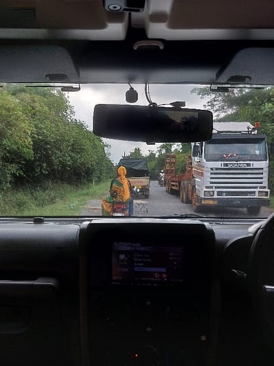 The image depicts a road scene in Kisanga, Tanzania, viewed from inside a vehicle. The foreground shows the interior of the vehicle, including a dark dashboard with a central infotainment screen displaying "BLUETOOTH Audio" and music details, a rearview mirror, and an attached dashcam. Outside the windshield, an overcast sky is visible above a narrow road bordered by dense green foliage on both sides. On the road, traffic is queued. A person, viewed from behind, wears a yellow head covering and a vibrant, multi-colored garment (yellow, green, blue patterns) and stands near a smaller, dark-colored truck with a canvas bed cover, partially revealing "VITA" on its side. Further down the road, a motorcycle with a rider is visible. To the right, a large white SCANIA truck with a flatbed trailer is prominent, displaying a Tanzanian license plate (e.g., T 882 DPU). Additional vehicles extend down the road, indicating a traffic halt or slow movement.