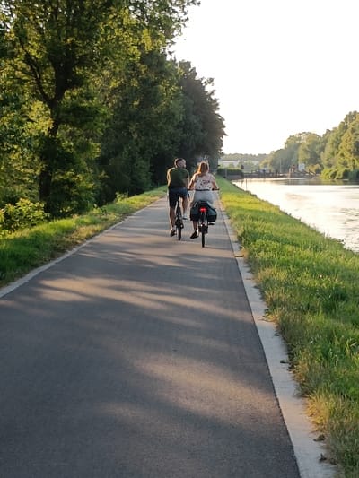 In the city of Haacht, Belgium, a paved pathway runs alongside a tranquil canal. The path is lined with lush green grass and mature trees casting dappled shadows. Two individuals are cycling away from the viewer on the pathway. The person in front, on the left, is a male wearing a green t-shirt and dark shorts. He is on a black bicycle. The person behind him, on the right, is a female wearing a white patterned top and light-colored shorts. She is also on a black bicycle, which has panniers attached to the rear rack. The canal reflects the bright sky, and distant structures, possibly a bridge or lock system, are visible across the water. The scene suggests a peaceful recreational activity in a natural setting.