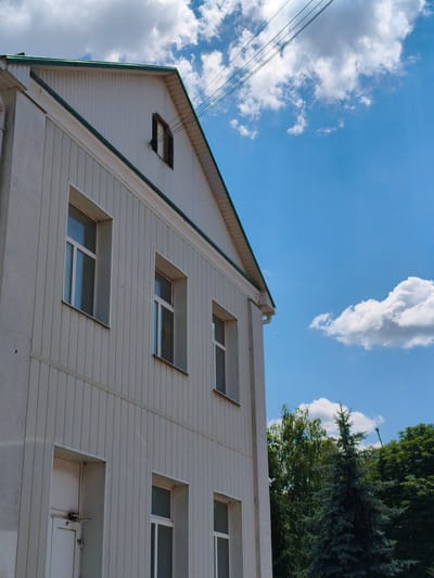 This is a medium-angle, full-shot, outdoor, daytime photograph taken in Velyka Bahachka, Ukraine. The image features the side of a two-story building, possibly an apartment complex or administrative building, clad in vertical white siding. The building has several white-framed windows with clear glass, some with internal grilles. On the upper level, there is a dormer window with dark shutters. A white door with a metal handle is visible on the ground floor. The building's roof is white with a green trim. Power lines are visible against a bright blue sky with scattered white clouds. In the background, lush green trees and a dark green fir tree are present. The scene is devoid of people.