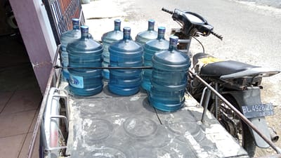 The image depicts a sidecar motorcycle loaded with several large, clear plastic water jugs, positioned in front of a building with a pinkish-purple wall and a metal fence. The motorcycle, with a black seat that appears worn and torn in places, is parked on an asphalt surface. A license plate on the rear fender reads "BL. 4928. ZD 08.20". The water jugs are arranged on the flatbed of the sidecar and in front of it, with at least seven visible. The scene appears to be a delivery or transport of bottled water. The environment suggests a sunny day in a residential or commercial area, possibly in Geulangganggampong, Indonesia, given the context provided.