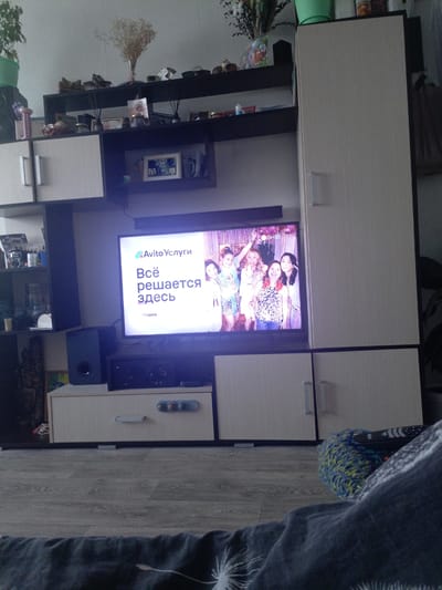 This media file depicts a living room scene in Podporozhye, Russia. The focal point is a large television screen displaying an advertisement for "Avito Услуги," a Russian online classifieds platform, with the slogan "Всё решается здесь" (Everything is solved here) and "Уборка" (Cleaning). The advertisement features a group of four young women smiling and posing for a photo.

The television is part of a modern entertainment center, which includes shelves with various decorative items, such as dried flowers in a vase, framed pictures, and other knick-knacks. There are also speakers and what appears to be a DVD player or receiver beneath the television. The entertainment center is made of light-colored wood with dark accents.

To the right of the television is a tall, light-colored cabinet with a single door and a handle. To the left of the television, a dark speaker is visible, along with some shelves containing items like a picture frame and a small plant. The floor is covered with light-colored laminate flooring.

In the foreground, a person's legs are visible, covered by a dark blanket and a colorful knitted sock. A remote control rests on the blanket. The overall impression is one of a cozy and lived-in domestic space. The lighting suggests it is daytime, but the exact time is not discernible. There are no indications of the weather. The emotion conveyed by the people on the screen is cheerful and positive.