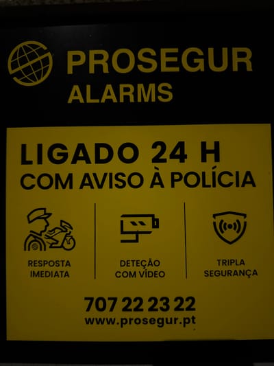 The image shows a PROSEGUR alarms sign, with the company logo on the left. To the right, the sign indicates "LIGADO 24 H COM AVISO À POLÍCIA," which translates to "CONNECTED 24 H WITH POLICE NOTICE." It also displays icons and text indicating "TRIPLA SEGURANÇA" (Triple Security), "DETEÇÃO COM VÍDEO" (Video Detection), and "RESPOSTA IMEDIATA" (Immediate Response). A phone number, 707 22 23 22, and website, www.prosegur.pt, are also printed on the sign.