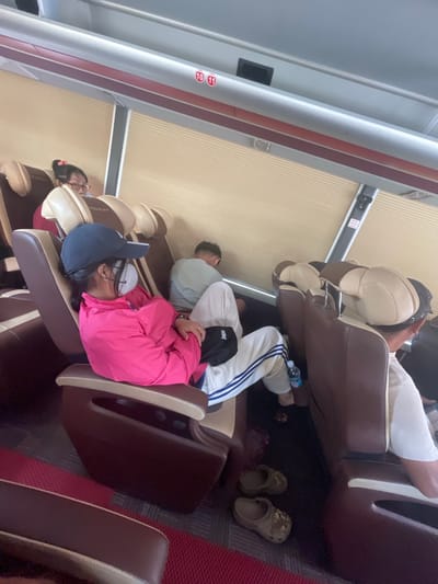 The image depicts the interior of a bus with reclining seats. In the foreground, a young woman wearing a pink jacket and a blue baseball cap is seated, facing away from the camera. She is wearing a white face mask and her arms are crossed. Next to her, to the right, another passenger is seated, also facing away from the camera. Further back, a person with glasses is visible in a seat. The bus interior features beige blinds, dark brown seats with beige headrests, and a red carpeted floor. A water bottle is on the floor next to the woman in the pink jacket, along with a pair of brown clogs. The bus appears to be stopped or parked. The sign visible in the upper part of the image indicates seat numbers 10 and 11. The location in the city of Gò Dầu, Vietnam, is noted.
