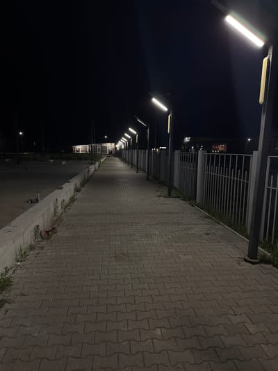 This is a nighttime, eye-level, wide shot of a paved pedestrian walkway in Chuguyevka, Russia. The walkway is constructed of grey interlocking pavers and extends into the distance, illuminated by a series of modern, linear streetlights mounted on dark poles. To the right of the walkway is a metal fence with vertical bars, and beyond that, faint lights suggest buildings or structures. To the left of the walkway is a low concrete barrier, with patches of sparse grass and vegetation growing along its base. Beyond the barrier, the ground appears to be unpaved and dark, possibly a parking lot or undeveloped area. In the far distance, there is a building with illuminated windows, and a few figures are visible near the entrance. The overall scene is quiet and somewhat desolate, with the artificial light casting long shadows.