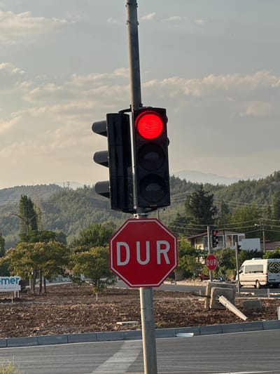 A red traffic light is illuminated above a large, red, octagonal stop sign with "DUR" written in white letters in Seydikemer, Turkey. A traffic light pole is visible to the right of the stop sign. Behind the stop sign, there is a smaller red stop sign with "DUR" written on it. The scene is outdoors, with a paved road in the foreground and a dirt lot behind it. Trees and a hill covered in evergreen trees are visible in the background, with power lines and distant mountains. A white van with "OKUL TASITI" written on it is parked to the right of the stop sign. The sky is partly cloudy.