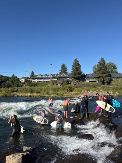 The image depicts an outdoor scene at the Bend Whitewater Park on the Deschutes River in Bend, Oregon, United States, under a clear blue sky. The foreground and mid-ground feature the river with a prominent whitewater wave on the left and a rocky, turbulent section on the right.
Approximately 11 individuals are present, engaged in or observing river surfing activities. In the mid-ground, a shirtless male in blue shorts is actively riding a white surfboard on a standing wave. To his left, another individual in a dark wetsuit holds a partially submerged pink and white surfboard while standing in the water. Further right, a male in a camouflage-patterned top stands in the water with a white surfboard in front of him.
On the right side, a group of approximately seven individuals are positioned on or near dark, wet rocks at the river's edge. This group includes shirtless males, individuals in dark full-body wetsuits, and others holding various river surfboards, including pink, light blue, and black boards with white undersides. One individual in a dark wetsuit wears an orange helmet.
The background across the river features a vegetated slope leading up to a multi-story building with distinctive gabled roofs. Tall evergreen trees and utility poles are also visible on the distant riverbank. The overall environment suggests a recreational area for river sports.