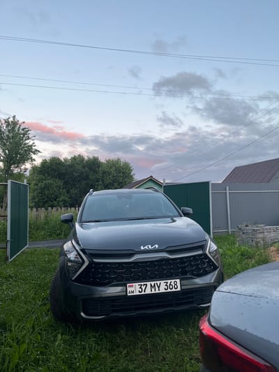 In the village of Klobushnevo, Russia, a dark gray Kia Sportage is parked on a grassy area in front of a wooden fence and a green gate. The car's license plate reads "37 MY 368". In the background, there are trees and houses. The sky is cloudy with some pink hues, suggesting it is either dawn or dusk. Another car is partially visible in the foreground, to the right of the Kia Sportage.