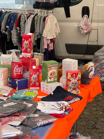 This image captures an outdoor market stall in Vila Nova de Cerveira, Portugal. The scene primarily features merchandise displayed on a table and a clothing rack.
In the foreground, a table draped with an orange tablecloth presents a variety of items. Prominently featured are multiple red boxes labeled "POP MART," showcasing a white, bunny-like character, with some boxes specifically indicating a "McDonald's" collaboration. Green boxes marked "THE MONSTERS VINYL FACE" with a similar creature in a monster costume are also visible. A large, red Coca-Cola can replica stands among the boxed items. Various other light-colored boxes are stacked. Folded t-shirts, some in clear plastic packaging, are spread across the table; designs include "SPIDER-MAN" on black shirts, and other cartoon characters on white and green shirts. A gray, plush, bunny-eared toy is positioned on the right side of the table.
Immediately behind the table, a metal clothing rack is laden with numerous children's garments, primarily long-sleeved tops and sweaters, hung on white plastic hangers. In the background, a white commercial van is parked, with a white plastic bag hanging from its side door handle. To the right of the van, a large, dark, bundled object is visible on the ground. The ground surface beneath the stall and van is paved with dark-colored cobblestones. No individuals are present in the image.