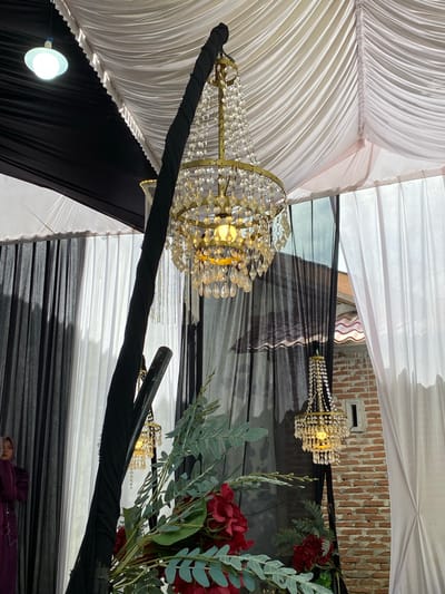 The image depicts an event space decorated with draped fabrics, chandeliers, and floral arrangements. The ceiling features black and white draped fabric sections, meeting diagonally across the upper left. A circular white downlight is visible against the black ceiling drape on the upper left. A large, illuminated gold-toned chandelier adorned with clear crystal beads and drops hangs prominently in the upper center. A thick black fabric-wrapped support pole is positioned diagonally, extending from the upper center to the lower left. Two additional, smaller gold-toned chandeliers with crystal accents are visible: one partially obscured behind the pole on the left, and another illuminated on the lower right. Dark sheer drapes frame the left and central areas, contrasting with white opaque drapes on the right. On the far left, a woman wearing a purple dress and hijab is partially visible, facing leftwards. In the lower center-right, a floral arrangement featuring prominent deep red flowers and green foliage is visible. An outdoor view through the right drapes reveals a textured brick wall with a small rectangular aperture and a curved terracotta-tiled roof section. The scene is located in Matangbaroh, Indonesia.