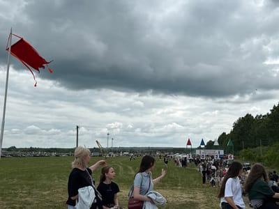 The image depicts an outdoor event, possibly a festival or fair, set in a wide, grassy field under a cloudy sky. A large red flag on a pole flutters in the wind in the foreground. Several people, including women and children, are visible, some looking towards the distance and others interacting with each other. In the middle ground, a crowd of people is walking along a path, with tents and structures suggesting vendor stalls or event areas. In the background, trees form a natural border, and there are signs with numbers and text, one of which appears to be in Russian. The overall scene suggests a lively public gathering in an open, rural or park-like setting. The location is stated as Narkeevo, Russia.