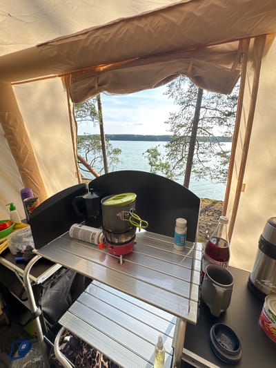 The image shows a campsite setup with cooking equipment and supplies laid out on a portable table in front of a tent. The scene is set by a body of water, likely a lake, bordered by trees, suggesting a natural outdoor environment in the vicinity of Priladozhskoe, Russia.
On the table, there is a portable stove with a pot on top, a moka pot, a fuel canister, a bottle of dark red liquid, a metal mug, and a small container. To the left, there is a green bowl with red chopped vegetables, a bottle of soap or sanitizer, and a rolled-up towel. A thermos is also visible on the right side of the table, along with a can of food. The table itself is made of a silver, slatted material. The tent fabric is visible at the top and sides of the frame, in a light beige color.
The background reveals a serene lake with calm water and a distant shoreline lined with trees. The sky is clear with some light clouds. The overall impression is one of outdoor recreation and self-sufficiency in a natural setting. No people are present in the image.