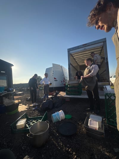 The image captures an outdoor commercial or service yard in Northallerton, United Kingdom, under a clear, bright sky casting long shadows. The ground surface is gravel.
Five individuals are visible. In the foreground on the right, a man with curly hair and sunglasses resting on his head looks downward towards the scene. Center-right, a man in a light-colored shirt and dark apron stands in front of a large white box truck with its rear door open, holding a metal tray above a stack of green plastic crates. Two additional men are inside the truck's cargo area; one is bent over, handling crates, while the other stands further inside. Behind them, a white panel van, branded "NORTHALLERTON / YORKSHIRE FOOD CO" and bearing a partial UK license plate beginning "YZ", is parked. To the left of the box truck, a woman in a white shirt and dark apron stands with her hands on her hips, holding a yellow cup. Further left, adjacent to a dark-colored shed or building, another woman in dark attire stands partially facing away from the viewer.
The ground in the foreground is occupied by various items, including multiple green plastic crates, three black refuse bags, a large metal cooking pot, a light blue plastic bucket lying on its side with a green lid, a black circular pan, and stacked white plastic food containers. The overall scene suggests a loading, unloading, or clean-up operation associated with a food service enterprise.