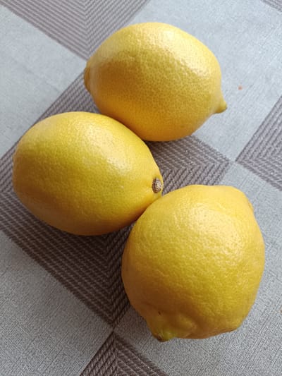 Three bright yellow lemons are arranged on a grey and white checkered tablecloth. One lemon is positioned at the top right of the frame, another to the left, and the third at the bottom center. The tablecloth has a geometric pattern of diagonal stripes. The lemons have textured skins, typical of citrus fruit. There is a small orange fleck visible on the tablecloth to the right of the top lemon.