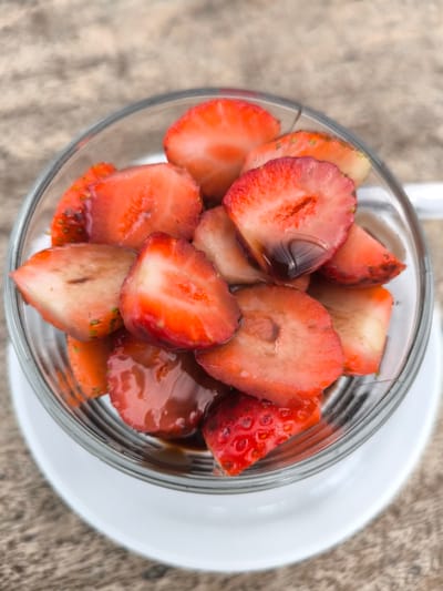 A close-up, overhead shot depicts a clear glass containing sliced strawberries with a dark, syrupy liquid. The glass sits on a white saucer. The backdrop shows a blurred, neutral-toned surface. The location is Candikuning, Indonesia.