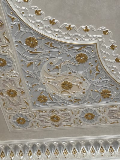 This image displays a close-up of intricate architectural ornamentation, likely on a wall or ceiling. The decorative elements are rendered in relief and feature a repeating pattern of floral motifs and swirling vines. The color palette is predominantly white and pale gray, with accents of gold. Small, golden studs or beads are interspersed throughout the design, adding further detail. The patterns are complex and appear to be executed with precision. There is no indication of people, a specific activity, time of day, weather, or emotions. The location context suggests Urgench, Uzbekistan, which may imply a style of Islamic art or architecture. The image does not contain any visible text.