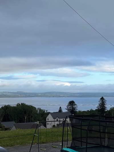The image depicts a scenic view of Mountcharles, Ireland, likely taken in the late afternoon or early evening, given the soft, diffused light. The scene is dominated by a vast expanse of water, possibly a bay or a sea, stretching to the horizon. Wispy clouds drift across a pale blue sky, creating a serene and slightly dramatic atmosphere.
In the foreground, a trampoline and a swing set are visible, suggesting a domestic setting, perhaps a backyard or garden. These objects are placed on a grassy area, with a paved path or driveway to the right. Several houses are scattered amongst the trees, some with dark tiled roofs, hinting at a rural or semi-rural environment. The houses appear modern in style.
The landscape beyond the water is gently rolling, with patches of green fields and some signs of human habitation, possibly small villages or isolated farms. A few wind turbines can be spotted in the distance, indicating the presence of modern infrastructure in the rural setting. Tall evergreen trees stand out against the lighter backdrop of the sky and water, adding depth to the composition.
The overall mood is peaceful and tranquil, evoking a sense of calm and natural beauty. There is no visible text in the image.