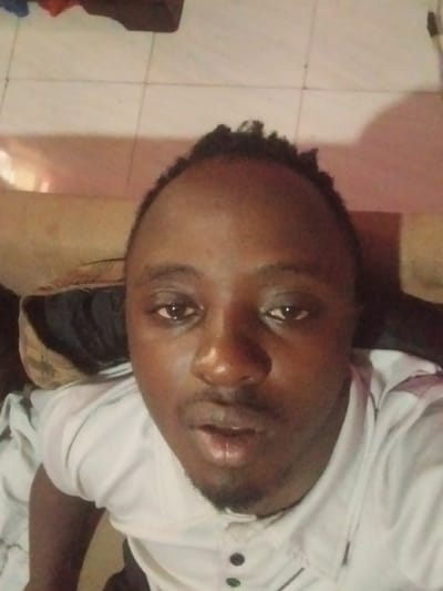 This scene, located in Riabai ward, Kenya, depicts a single adult male in a close-up, high-angle shot, likely a selfie. The male has a dark complexion, short dark textured hair, and a slight goatee. He wears a light-colored, possibly white or light grey, collared shirt with a partial button placket featuring three dark buttons. His eyes appear slightly red or moist, and his mouth is slightly open. The background is an indoor setting with elements out of focus, including light-colored tiled flooring with dark grout lines visible in the upper portion of the frame, and a blurred light brown/beige upholstered surface (possibly a cushion or sofa arm) to the subject's upper left. Indistinct dark and reddish objects are also visible in the background. The lighting is artificial and somewhat dim, casting a subtle shadow on the subject's right cheek.