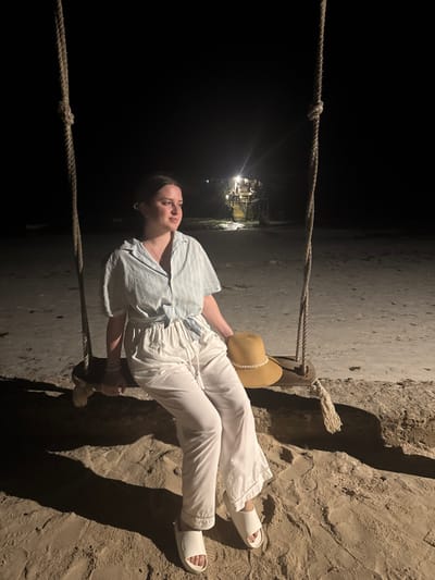 A woman in a light blue striped shirt and white pants sits on a swing, holding a straw hat. She's wearing white sandals. The swing is made of rope, and the backdrop appears to be a sandy beach at night. A dimly lit structure is visible in the distance. The image is slightly rotated.