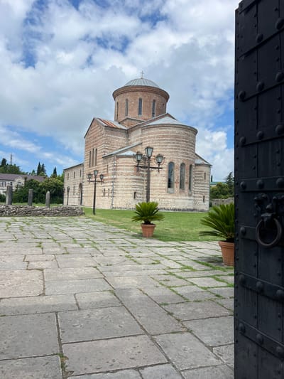 A low-angle, exterior, full shot shows the partially open, large, black metal gate of an ancient brick building in Pitsunda, Abkhazia. The gate's surface is textured with raised circular rivets and horizontal bands, and a large ring handle is visible. Beyond the gate, a vast, grey stone courtyard stretches towards a majestic, ancient brick church with a domed roof topped by a cross. The church exhibits a distinct architectural style with alternating bands of red brick and lighter stone. In front of the church, a manicured green lawn hosts two potted palm plants and several old-fashioned lampposts. To the left of the courtyard, ancient stone pillars and a low stone wall mark the remnants of a previous structure. In the background, a cluster of trees and a building with a red roof can be seen under a bright, cloudy sky.