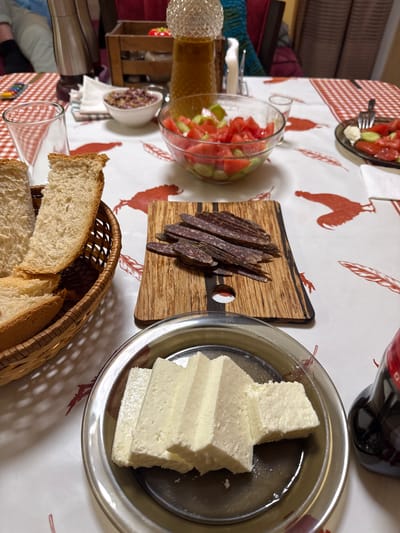 The image shows a table laden with food. There's a basket of sliced bread, a wooden board with sliced cured meat, a metal plate with blocks of white cheese, a glass bowl of sliced tomatoes and cucumbers, a small bowl of olives, a bottle of liquid, and glasses. The tablecloth is white with red rooster motifs. The scene potentially represents a meal setting. It is not possible to verify the location.