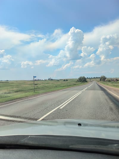 The image depicts a view from inside a vehicle traveling on a paved road through a rural landscape. The road has a double white line marking the center. On the left side of the road, there is a grassy field with a few scattered trees and a blue signpost with white lettering indicating "487". In the distance, there are farm buildings and structures. The sky is blue with fluffy white cumulus clouds. The foreground shows the dashboard of the vehicle. The scene appears to be a sunny day in a rural area of Старокутлумбетьево, Russia, suggesting a journey through the countryside.