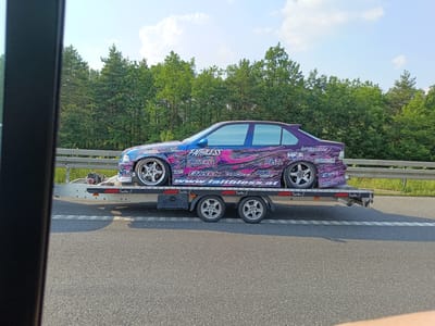 The image shows a custom-painted blue and purple BMW sedan on a trailer, likely a BMW E36. The car features a vibrant livery with stylized graphics and sponsor logos. Visible text includes "FAIRLESS," "DRIFTSTROCE," "SSR," "bimare," "WWORK," "HKS," "VALINO," "BLTZ," "326power," and "www.faithless.at." The trailer itself is labeled "JAGRI Trailer" multiple times.

The vehicle is being transported on a two-axle trailer, which is being towed on a paved road. The scene is set outdoors during the daytime, with a clear blue sky and fluffy white clouds visible. A line of green trees forms the background, suggesting a location in or near a forested area. A metal guardrail is visible between the road and the trees. The overall impression is of a car being transported, possibly to or from a car show, drift event, or race. The lighting and clear sky indicate fair weather. The image appears to have been taken from inside another vehicle, with a dark frame on the left side of the image indicating a window or door.