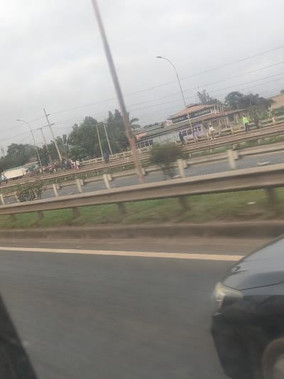 The image displays an outdoor, daytime scene in Kiuu ward, Kenya, captured from the interior of a moving vehicle. The immediate foreground features the front right quarter of a dark-colored vehicle, including its headlight and partial hood, positioned on a multi-lane asphalt roadway marked with a white line. This roadway is separated by a series of parallel, metal guardrails from a grassy median or shoulder area.
Beyond the guardrails, a secondary road or shoulder is visible, populated by numerous pedestrians. On the left, a white commercial truck, possibly a box truck, is parked or moving slowly, with a cluster of individuals nearby. In the mid-ground, a two-story building with a distinctive purple lower section and a brown upper section is prominent. The building's facade clearly displays the text "SYNTAX ACADEMY" in white capital letters. Several pedestrians are observed walking or standing in front of this building, one of whom is distinctly wearing a bright green upper garment. The environment includes multiple utility poles, lampposts with overhead lines, and a variety of trees and bushes. The sky overhead is overcast, indicating diffused daylight conditions.