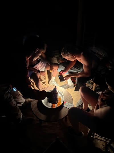 Four individuals are gathered around a low table in what appears to be an outdoor setting at night, illuminated by a single light source casting strong shadows. The environment is dark and appears to be rustic. On the table is a metal tray with small glasses containing an amber liquid, and a teapot or kettle from which liquid is being poured. One person, wearing a dark t-shirt and shorts, is seated on a stool and pouring the liquid. Another person, shirtless and wearing dark pants, is seated and looking at a smartphone. A third person, whose lower body is visible, is wearing shorts and white socks with sneakers, and is holding a yellow object. The fourth person's presence is suggested by their legs and arms in the foreground, also in darkness. A QR code is visible on a card on the table. The location is identified as Douar Ait Daoud, Morocco, which is a rural village.