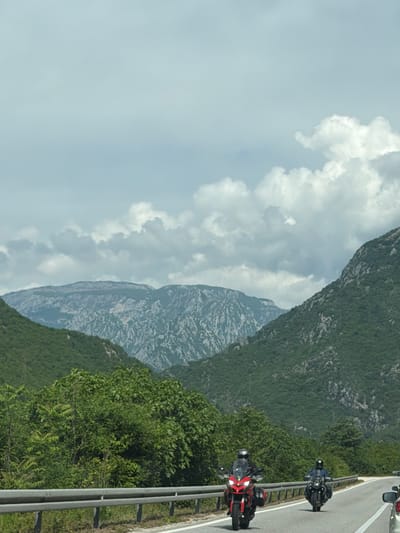 A scenic view shows a road with two motorcycles, a red one in the foreground and a black one further away. The road is bordered by a guardrail and lush green vegetation. A large mountain stretches into the distance, partially obscured by cloudy skies. The location is identified as Donji Jasenjani, Bosnia and Herzegovina.