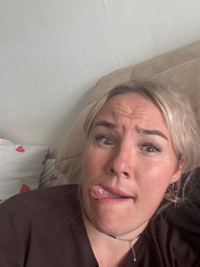 This is a selfie of a blonde woman in a dark brown top, sticking her tongue out. She has on false eyelashes and a necklace. The background appears to be a bed with a pillow and a white duvet with red hearts. The image was taken in Pozdyshki, Russia.