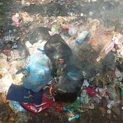 The image displays an open dump site containing mixed refuse in Pasir Muncang, Indonesia. Two prominent black plastic refuse bags are centrally located; one exhibits active combustion with visible orange flames, producing significant white and gray smoke. Adjacent to the burning bag is a light blue plastic bag, filled with contents and adorned with colorful patterns. The surrounding debris comprises assorted household and commercial waste, including various plastic packaging, discarded cups, crumpled foils, organic matter, and textile fragments (notably a dark blue item with "Walking on Water" text and a red and white patterned fabric). Several blue plastic utensils are also visible on the dark earth ground, which serves as the base for the extensive waste pile. No individuals are present within the frame.