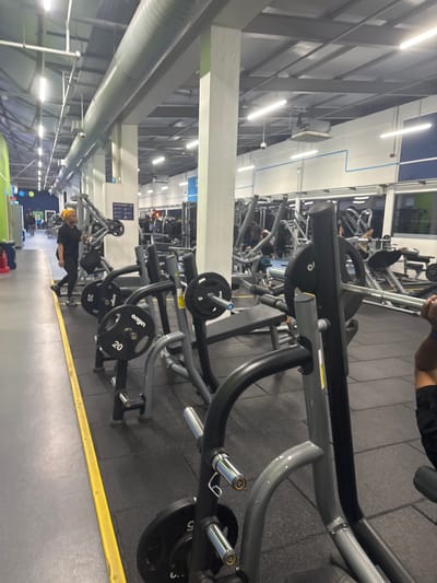 The image shows an indoor view of a gym, potentially located in Sheffield, United Kingdom. Gym equipment dominates the scene, including various weight machines with black weight plates labeled "origin" and numerical values such as "20" and "5."

A person wearing a yellow head covering and dark clothing is visible in the background using one of the machines. Other individuals are also present in the background. The floor is covered in dark gray rubber matting. Overhead lighting consists of multiple fluorescent tube lights. A large cylindrical duct runs along the ceiling on the left side of the image. Blue and white wall paneling are also visible.