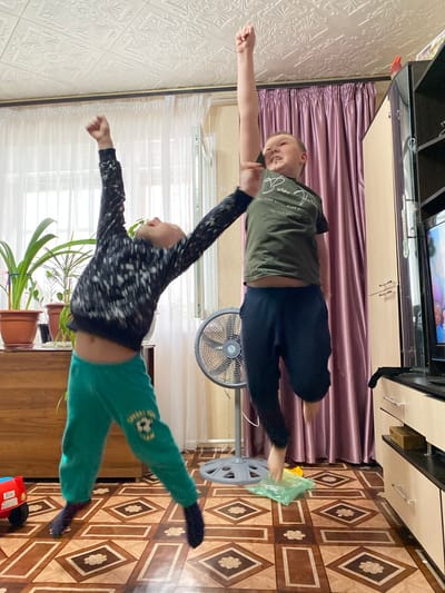 Two boys are lying on their stomachs, outstretched, in a room. One boy wears a green t-shirt and blue pants; the other wears a black and white long-sleeved shirt and green pants. A fan is in the background. Potted plants are visible in the lower part of the scene, and flooring with diamond-shaped patterns is visible on the right. A television stand with a TV is in the upper part of the scene. The room appears to be in a residential dwelling. There is no indication that the scene is located in the city of Bely Klyuch, Russia.