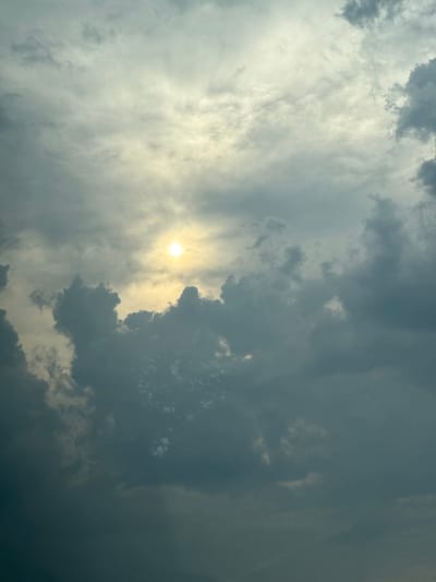This is a vertical, medium-shot photograph of a dramatic sky with the sun partially obscured by clouds. The sun appears as a bright, diffused circle of yellow light towards the center-left of the frame, casting a hazy glow through the surrounding clouds. The clouds themselves are a mixture of dark, stormy grays and lighter, wispy whites, creating a sense of depth and texture. Some of the darker clouds have a brooding, almost bruised appearance, while others are illuminated from behind by the sun, appearing translucent and ethereal. The overall impression is one of a turbulent sky, possibly hinting at an approaching storm or the aftermath of one. There are no people or objects visible, and the scene is solely focused on the atmospheric display. The lighting suggests it could be late afternoon or early morning, when the sun is lower in the sky and its rays are more likely to be diffused by cloud cover. There are no visible text elements in the image. The context provided indicates the location is Brashlen, Bulgaria.