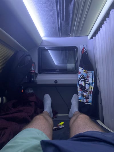 The image depicts an individual's legs and feet in a confined personal space, possibly a cabin or a small room. The person is wearing light grey socks and dark navy blue shorts. The space is dimly lit, with a bright light source visible on the left side, illuminating blinds or curtains.
Several objects are present in the scene. A dark-colored backpack is situated on the floor. Above it, a shelf holds a Coca-Cola bottle. To the right of the bottle, a black t-shirt with a colorful graphic print is laid out. A small screen or monitor is integrated into the wall unit. On a surface to the right, a yellow lighter and a small item with text are visible. A dark red blanket or piece of fabric is draped over a portion of the space, partially covering the backpack and extending towards the person's feet.
The overall impression is one of a private, possibly travel-related setting. The confined space and the presence of personal belongings suggest a temporary living or resting area. There are no clear indicators of the time of day or weather conditions. The lighting suggests it might be nighttime or an artificially lit interior. The visual cues of the blinds and the furniture suggest an indoor environment. No specific emotions are conveyed by the image.
Visible text includes "Coca-Cola" on the bottle. There is also text on the t-shirt, although it is partially obscured and difficult to read clearly. The yellow lighter also has some writing on it.