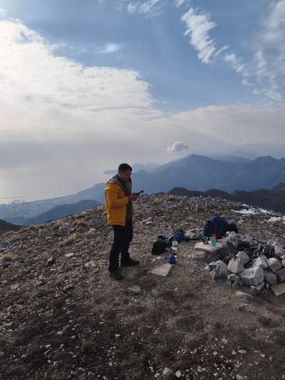 The image displays a male individual standing on a rocky mountain summit in Mali Mikulići, Montenegro. The man is clad in a yellow jacket, dark trousers, and dark footwear, looking down at a small dark electronic device held in his hands. To his right, two backpacks (one blue, one black) are placed on the ground next to a clear plastic water bottle, a blue water bottle, and three mugs (two green, one reddish) resting on a flat stone. Adjacent to these items is a circular arrangement of light-colored rocks. The ground is characterized by loose rocks and sparse, dry vegetation. In the background to the left, a large body of water, likely the Adriatic Sea, stretches to a populated coastline. Multiple layers of distant mountains are visible across the horizon, with some higher elevations exhibiting small patches of snow. The sky is partly cloudy with visible blue sections.