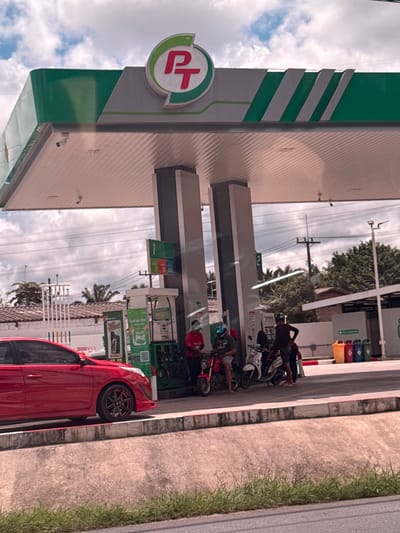The image depicts a gas station with a red car prominently parked in the foreground. The gas station features a distinctive green and white color scheme, with a large "PT" logo visible on the left side. Several motorcycles are parked near the pumps, with some people sitting on them, suggesting a brief stop or waiting period. The scene appears to be a typical day at a gas station in Ban Krasom, Thailand, with a mix of cloudy and clear skies overhead, indicating potentially mixed weather. The presence of tropical trees in the background further supports the Thai location. Some signs with text are visible at the gas station, though they are not clearly legible. The overall impression is of a busy, functional gas station with people and vehicles in transit.
