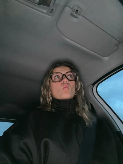 A person wearing glasses and a black jacket looks upward, puckering their lips, inside of a vehicle. The car's interior includes a gray ceiling, sun visor, and a cloudy sky visible through the window. The image appears to be taken at an angle, with the person positioned lower than the camera.