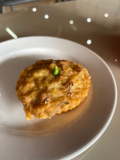 A single, round, golden-brown fritter, possibly a type of bakwan or perkedel, is presented on a white ceramic plate. A small, vibrant green chili pepper is skewered upright in the center of the fritter, serving as a garnish. The fritter has a slightly irregular, textured surface and appears to be made from a mixture of vegetables and batter, with visible flecks of darker ingredients within. The plate rests on a light-colored, reflective surface, possibly a table, where light glints and reflects in several small circles, suggesting an indoor setting with ambient lighting. The background is out of focus, showing muted tones and further reflections. The overall scene evokes a sense of simple, rustic cuisine. The location is the city of Bireuenmeunasahdayah, Indonesia.