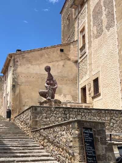 The image depicts a street scene in Pollença, Spain, featuring an outdoor sculpture of a seated, hunched figure, possibly a woman, positioned on a stone platform. The sculpture is made of a reddish-brown material and has a stylized, elongated form. Behind the sculpture, an aged, light-brown building with textured walls and visible stone construction rises. To the right, a larger, taller building with a light-colored stone facade, featuring a repeating pattern of small indentations, is visible. This building has several windows, some with barred exteriors.

In the foreground, a wide stone staircase ascends to the left, bordered by stone walls and metal handrails. On the right side of the staircase, a blackboard menu is propped against a stone pillar. The blackboard lists various food and drink items, including "Freshly Baked," "Ensiemada," "Croisan," "Coca de Patata," "Empanadas," "Tapas," "Calamares," "Tortilla de Patatas," "Croquetas," "Pimientos Padron," "Sangria," and "Copa Uno." A black trash bin is situated on the left side of the staircase. The sky is a clear, bright blue with a few wispy clouds. The overall scene suggests a warm, sunny day in a historic European town.