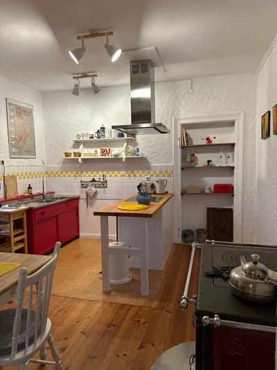 The image shows a kitchen in Ammeldingen bei Neuerburg, Germany. The kitchen features red cabinets under a stainless steel hood and backsplash tiles with yellow accents. A wooden countertop island with a white base is positioned in the center of the room. Shelving units are mounted on the wall and in a doorway, displaying various kitchenware and decorative items. A dark-colored stove with a silver teapot on top is visible in the foreground. Wooden flooring and a cork floor area are present. The room is illuminated by track lighting on the ceiling. No people are present in the scene.