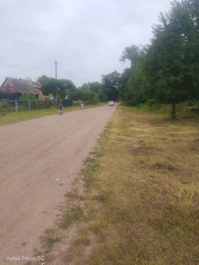 The image depicts a rural scene in Sestrynivka, Ukraine, under an overcast sky. A dirt road runs diagonally from the foreground into the distance, bordered on the left by a fence and a building with a dark roof. Beyond the fence, trees and some low-lying structures are visible. On the right side of the road, dry, yellowish grass is abundant. Two individuals are present: one, a young person on a bicycle, is in the middle ground on the left side of the road, facing away from the viewer. Further down the road, another person is walking, also facing away. A silver car is visible in the distance on the road. The overall impression is one of a quiet, everyday moment in a rural Ukrainian village.
