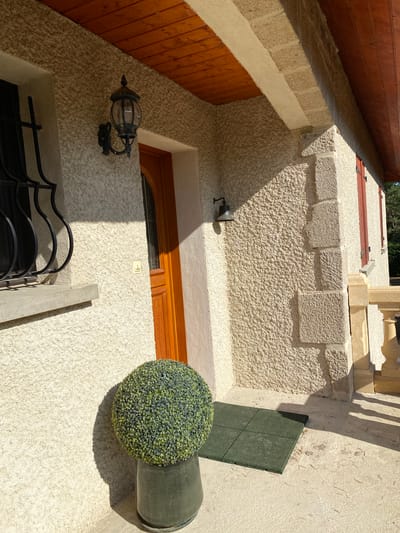 The image shows the exterior facade of a building, likely a house, in Courpière, France. The facade is constructed with rough-textured, light-colored plaster and features a wooden-framed door with a glass inset. Above the door, a dark metal lantern with glass panes is mounted on the wall. To the left of the door, a window is protected by decorative black wrought iron bars. A grey concrete windowsill is visible beneath the window. A spherical, green artificial topiary is placed in a dark green pot on the ground to the left of the door. In front of the door, a small, dark green mat composed of four square tiles lies on the concrete ground. The scene is bathed in sunlight, casting distinct shadows across the facade and the ground. The architectural style suggests a traditional or rustic aesthetic.