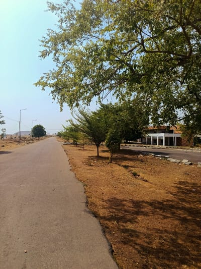 A road in Rafin Pa, Nigeria stretches into the distance, flanked by trees and arid ground. A low-rise building with a covered walkway is visible on the right. The sky is clear. There are no people visible.