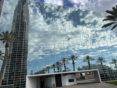 The image depicts the exterior of the Crystal Cathedral in Garden Grove, California, USA, a modern architectural marvel with a glass facade. The building's reflective surfaces mirror the bright blue sky filled with fluffy white clouds and the silhouettes of palm trees, creating a striking visual effect. In the foreground, the textured pavement leads to the entrance of the building. The scene is devoid of any apparent action or interaction among individuals, offering a serene and contemplative view of the structure.