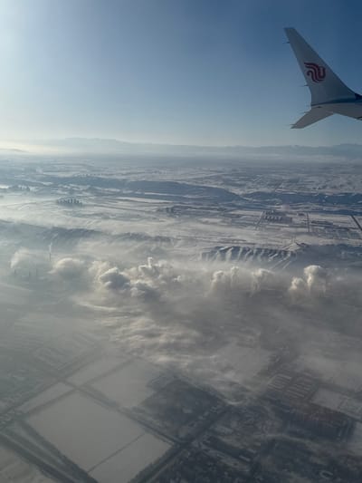 An aerial photograph depicts a winter landscape over or immediately adjacent to the city of Ürümqi, China. The upper right quadrant features the white wing and blue/red "swallow" logo of a China Eastern Airlines aircraft, indicating the viewpoint from an airplane. The ground below is extensively covered in snow, signifying winter conditions. The lower and mid-ground reveal significant industrial activity, characterized by numerous facilities with multiple smokestacks emitting substantial plumes of white and grey exhaust. These plumes coalesce to form a dense, low-lying layer of atmospheric haze or smog that obscures visibility across the terrain. Beneath this haze, structured residential or industrial blocks and what appear to be snow-covered agricultural fields are visible in a grid pattern. Beyond the immediate industrial zone, snow-covered hills with distinct ridges rise, and further in the distance, additional snow-dusted mountains and plains extend towards a hazy horizon. The sky above the haze is clear and blue. No individual persons or their direct interactions are discernible from this altitude.