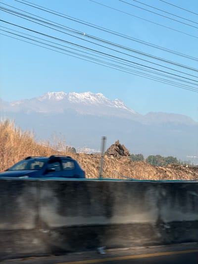 The image presents a daytime view captured from a moving vehicle in the vicinity of Santa María Zacatepec, Mexico. In the immediate foreground, a concrete highway barrier runs horizontally across the lower third of the frame, showing motion blur, with a yellow road line visible on the asphalt below. A dark blue hatchback vehicle, equipped with roof rails, is partially visible above the barrier in the left-center, also blurred due to motion. Behind the barrier, the terrain consists of dry, brown earth with scattered sparse vegetation, leading to a small, dark, irregular rock formation on the right side of the mid-ground. Further in the distance, green trees partially obscure a cluster of white and light-colored structures, indicative of a town or city, with a prominent street light pole visible. The background is dominated by a large mountain range with snow-capped peaks, partially veiled by atmospheric haze. The sky above is clear blue, and a faint crescent moon is discernible in the upper right quadrant. Multiple parallel black utility lines traverse the upper portion of the sky diagonally.